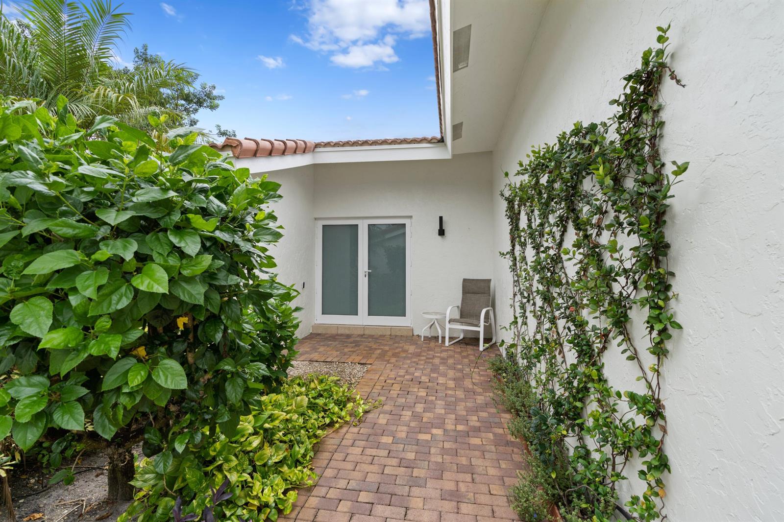 A brick patio leads to a glass-paneled door beside a wall covered in climbing vines — 925 Foxpointe Circle, Delray Beach FL 33445, DELRAY BEACH, FL