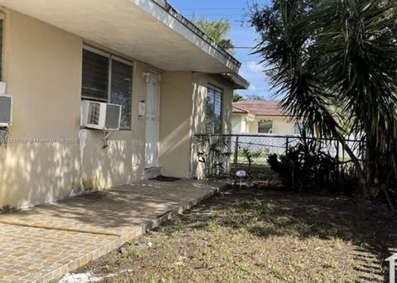 A beige single-story house exterior features a tiled porch, a window air conditioning unit, and landscaping — 409 SW 4th Ter, Hallandale Beach FL 33009, HALLANDALE BEACH, FL