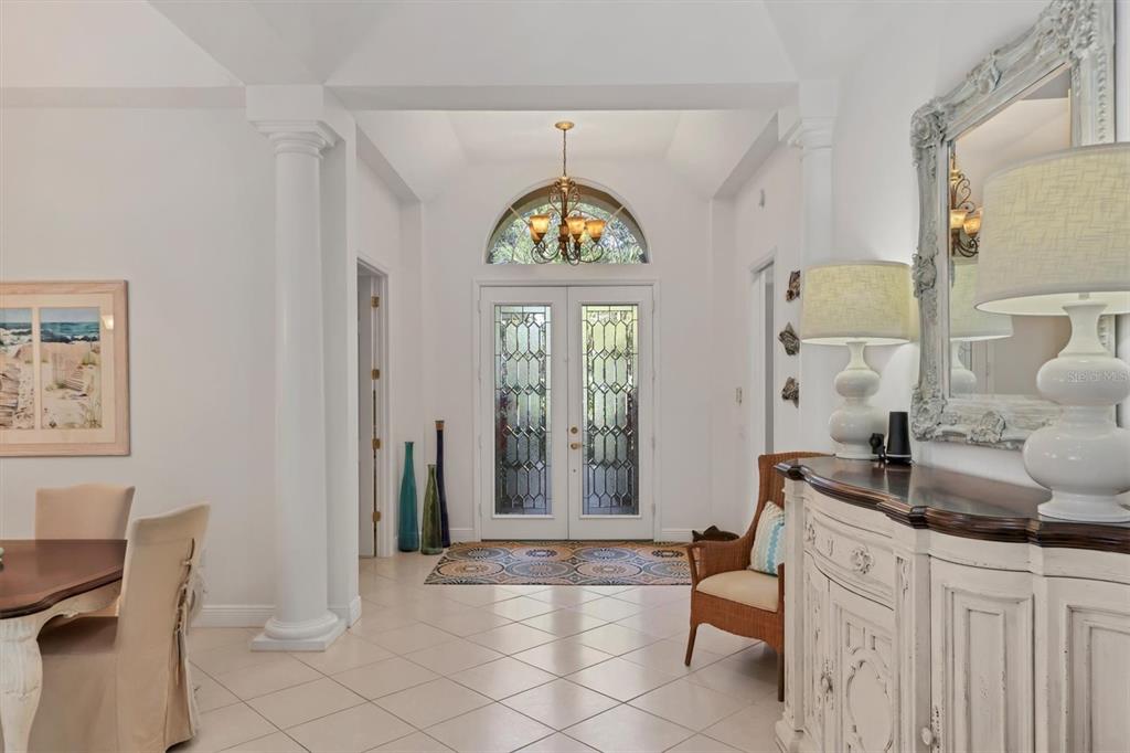A bright foyer features white columns, a decorative chandelier, glass-paneled double doors, and tile flooring — 8135 ABINGDON COURT, UNIVERSITY PARK, FL