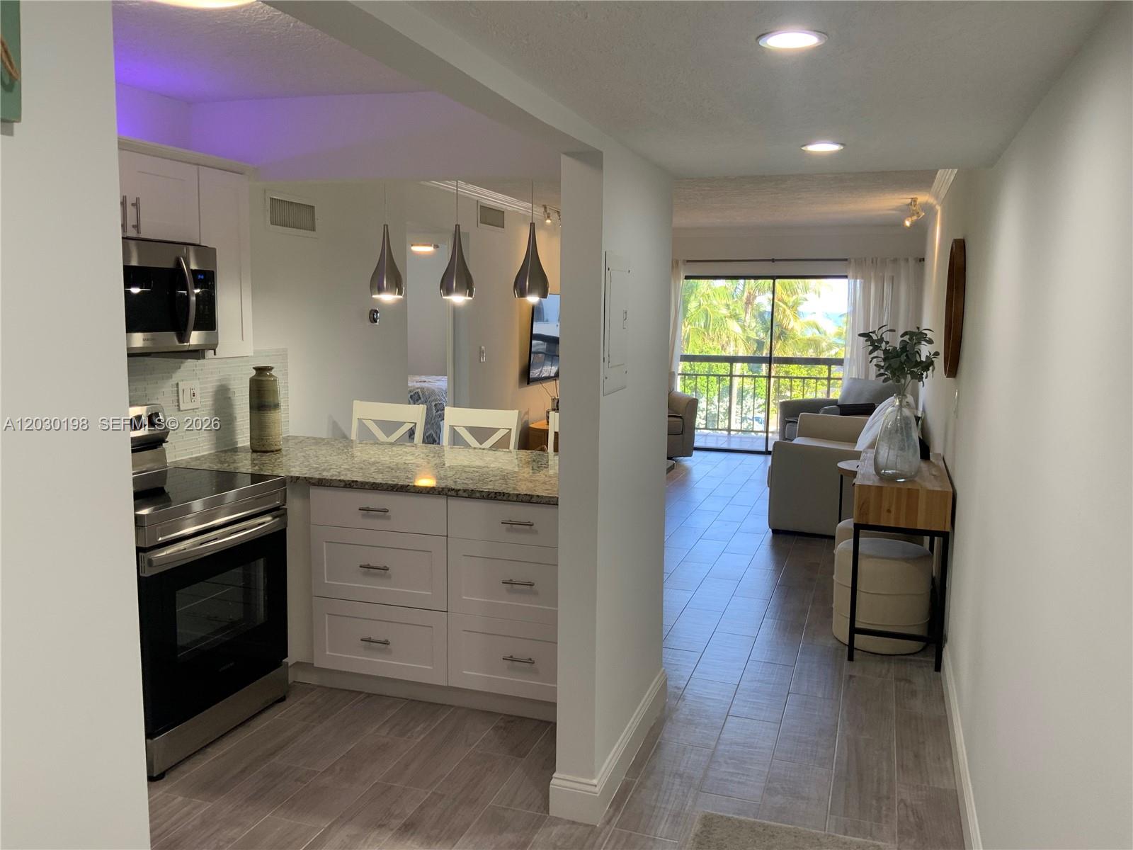 A kitchen with white cabinets and granite countertops opens into a living area with balcony — 6444 Collins Ave # A6, Miami Beach FL 33141, MIAMI BEACH, FL