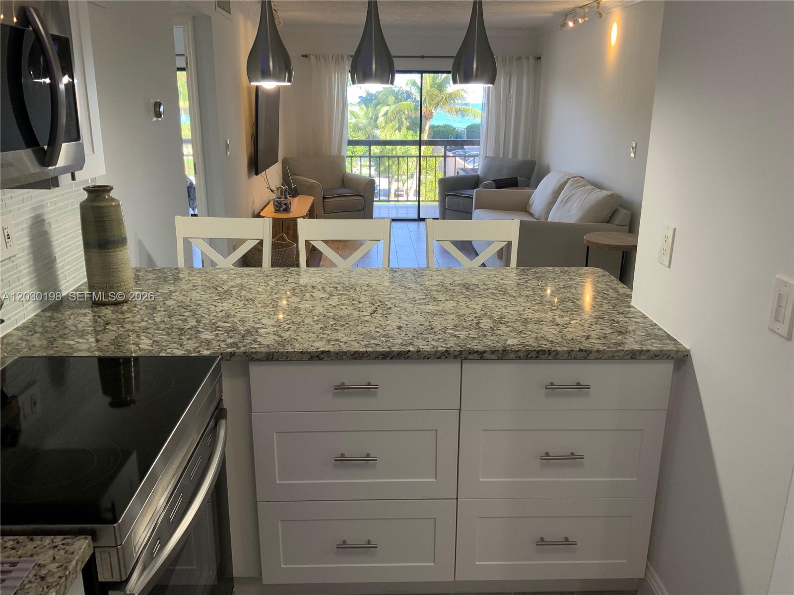 A kitchen with white cabinets and granite countertops overlooks a living area with balcony views — 6444 Collins Ave # A6, Miami Beach FL 33141, MIAMI BEACH, FL