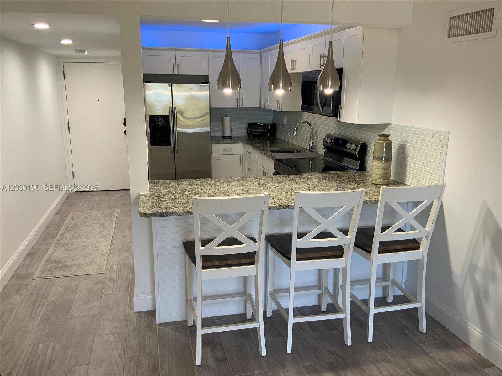 A kitchen features white cabinets, granite countertops, stainless steel appliances, and three bar stools — 6444 Collins Ave # A6, Miami Beach FL 33141, MIAMI BEACH, FL
