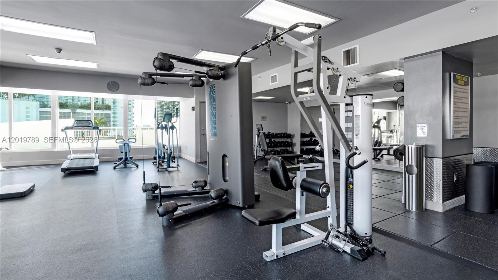 A fitness center features various exercise machines, rubber flooring, and large windows overlooking a city — 275 NE 18th st # 1606, Miami FL 33132, MIAMI, FL