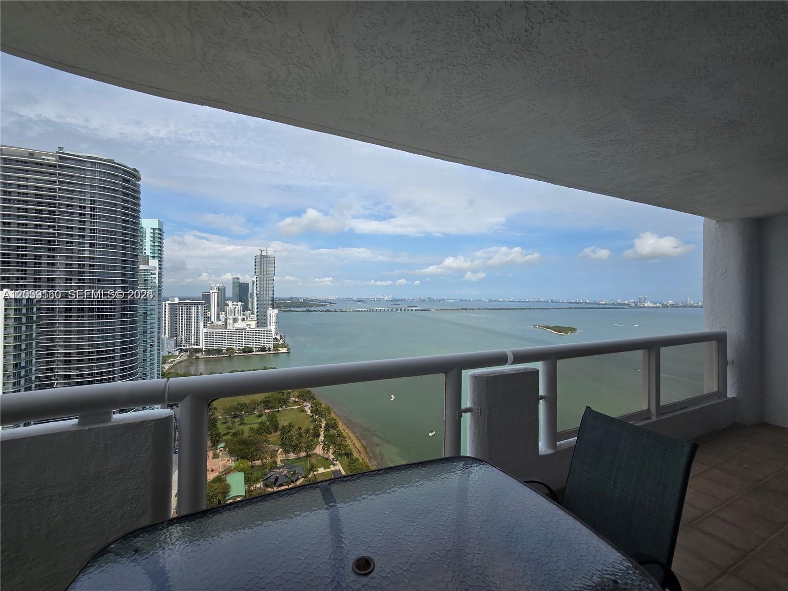 A balcony with a glass table and chair overlooks a bay, park, and city skyline — MIAMI, FL