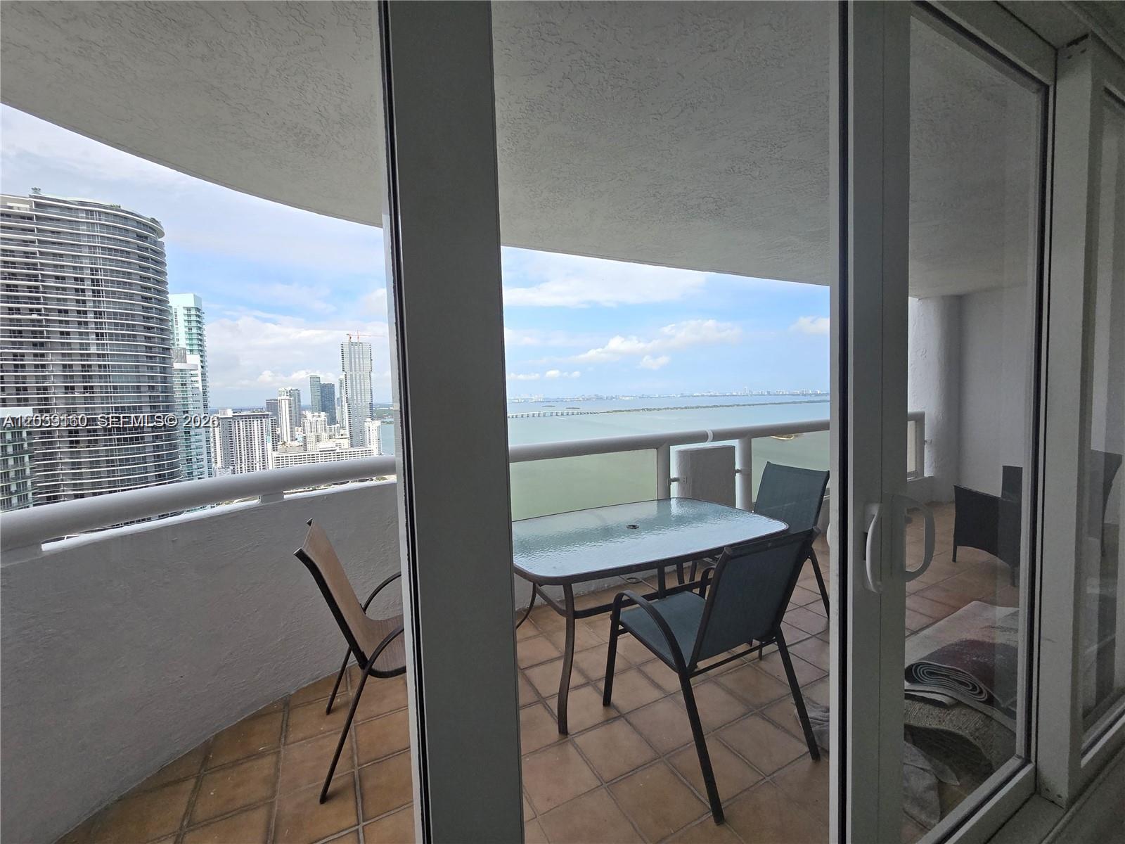 A balcony with a glass-topped table and chairs overlooks a city skyline and water — MIAMI, FL
