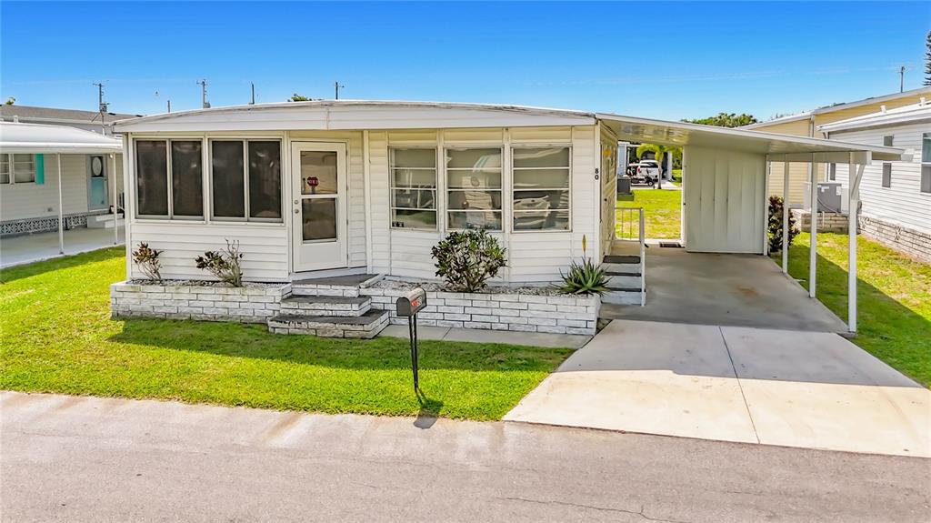 A white mobile home with a carport and a screened front porch sits on grass — 80 DEL RIO DRIVE, PALMETTO, FL