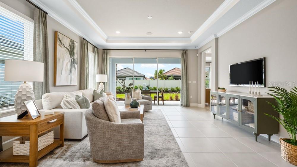 A living room features a tray ceiling, tile flooring, a television, and sliding glass doors — 3880 IMMACOLATA WAY, WESLEY CHAPEL, FL