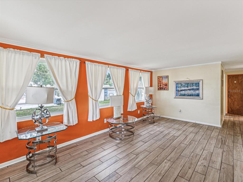 A living room features orange walls, wood-look tile flooring, three windows with curtains, and tables — 1940 SHADY COVE DRIVE, HOLIDAY, FL