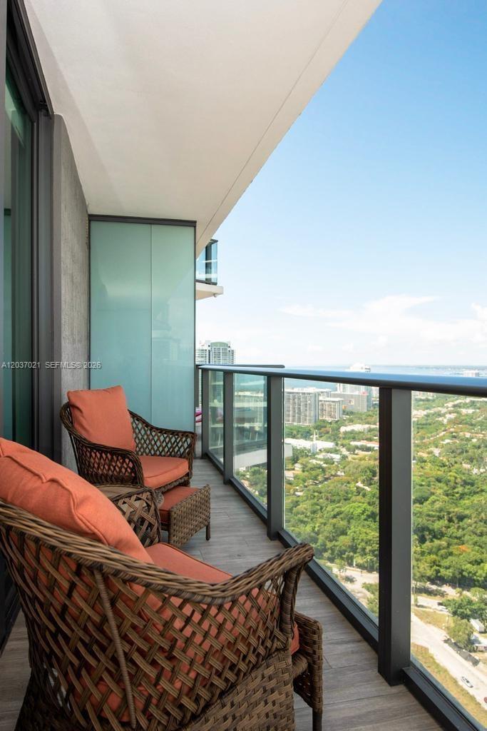 A high-rise balcony features wicker chairs with orange cushions overlooking a lush green landscape — 1300 S Miami Avenue # 3908, Miami FL 33130, MIAMI, FL