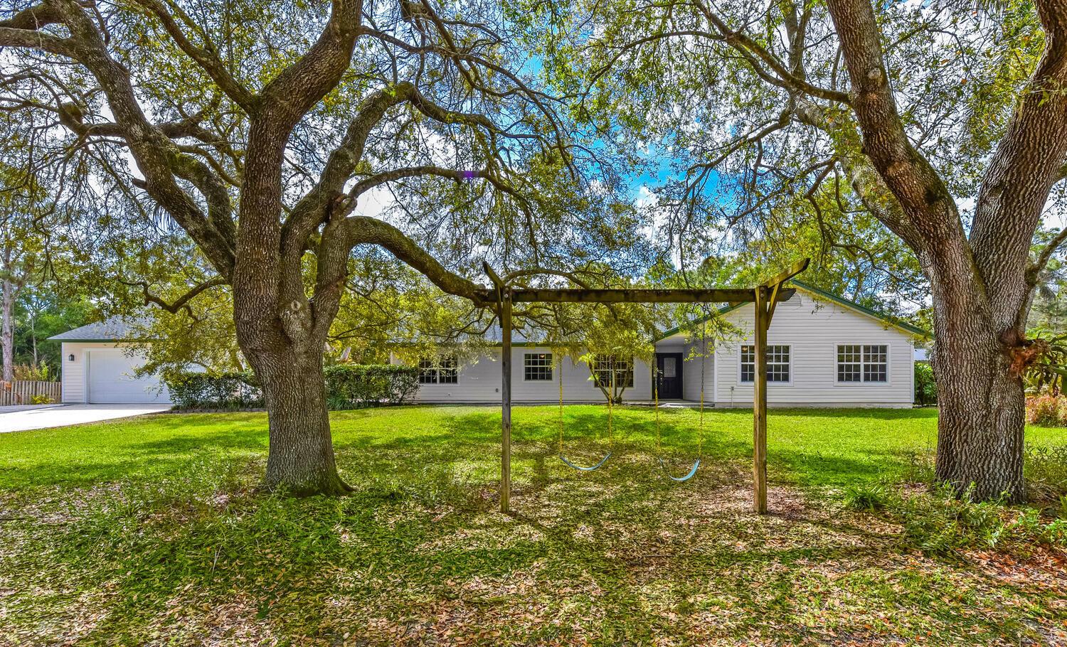 A white single-story house with a swing set in the front yard under large trees — 16244 E Downers Drive, Loxahatchee FL 33470, LOXAHATCHEE, FL