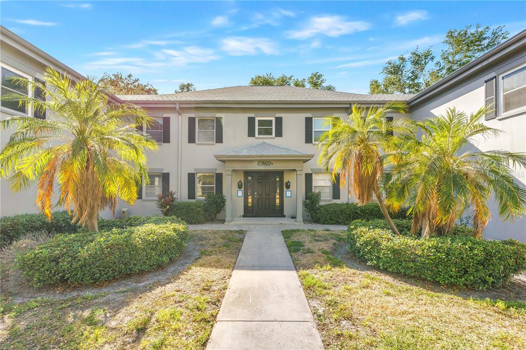 A two-story apartment building exterior features a central entrance, palm trees, and landscaped bushes — 6960A SUNSET DRIVE S UNIT 1B, SOUTH PASADENA, FL