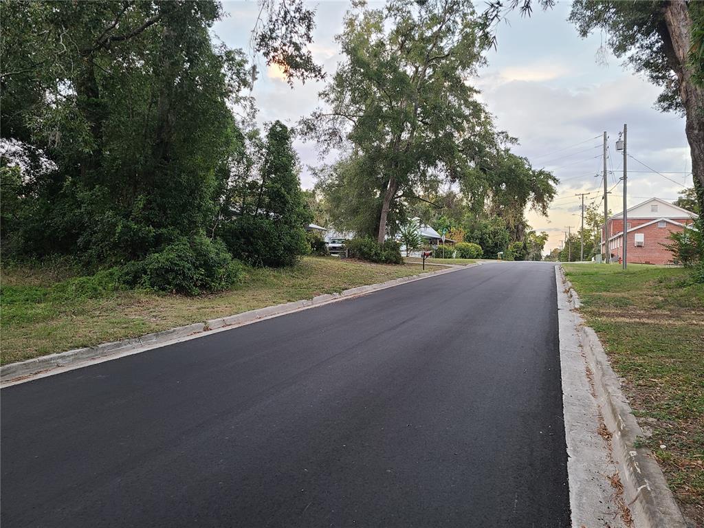 A paved residential street lined with mature trees and a brick building on the right — NW 183 PLACE, HIGH SPRINGS, FL