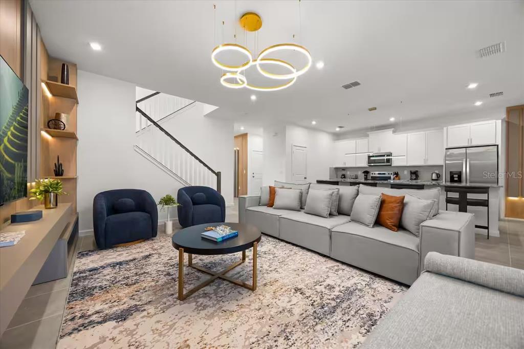 A modern open-concept living room features a grey sectional, two navy chairs, and a contemporary chandelier — 2337 SALTY WINDS WAY, KISSIMMEE, FL