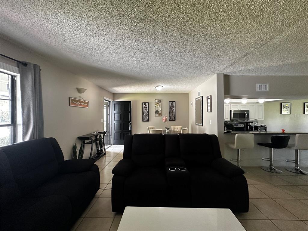 A living room features tile floors, a popcorn ceiling, and an open view to the kitchen — 5434 SADDLEBROOK WAY UNIT 3, WESLEY CHAPEL, FL