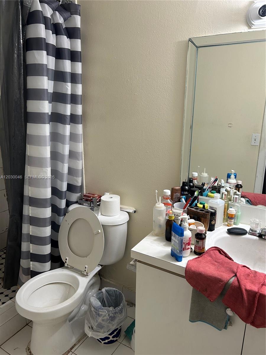 A bathroom features a toilet, a vanity with a cluttered countertop, and a striped shower curtain — 18051 NW 68th Ave # K104, Hialeah FL 33015, HIALEAH, FL
