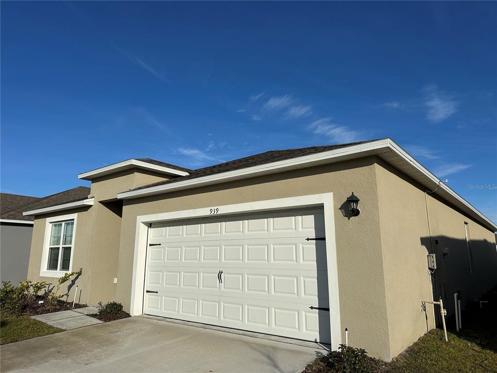 A tan single-story house exterior features a white two-car garage door and a side window — 939 SUN BURST ROAD, WINTER HAVEN, FL