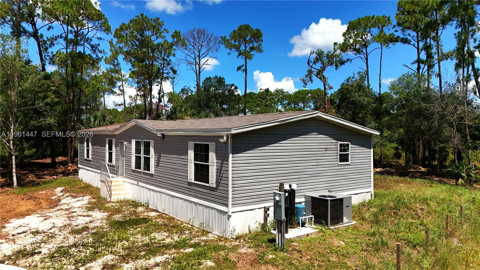A gray manufactured home sits on a dirt lot surrounded by pine trees — 881 WOODLAND BLVD # 0, Clewiston FL 33440, CLEWISTON, FL