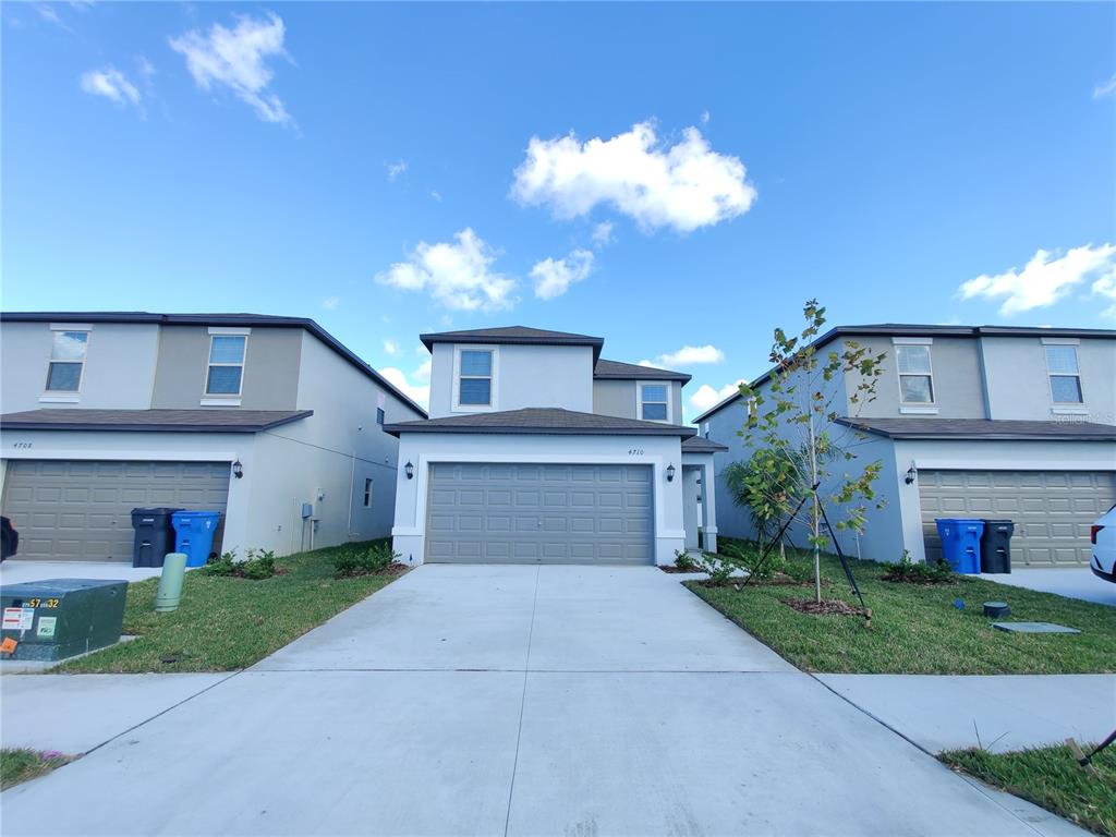 A three-unit residential townhouse complex with grey garages and light-colored stucco exteriors under blue skies — 4710 NORMAN OAK PLACE, TAMPA, FL