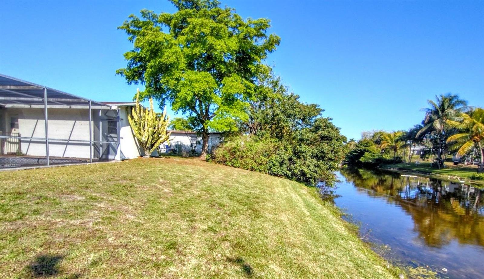 A grassy backyard slopes down toward a canal next to a house with a screened-in patio — 8528 Windsor Drive, Miramar FL 33025, MIRAMAR, FL
