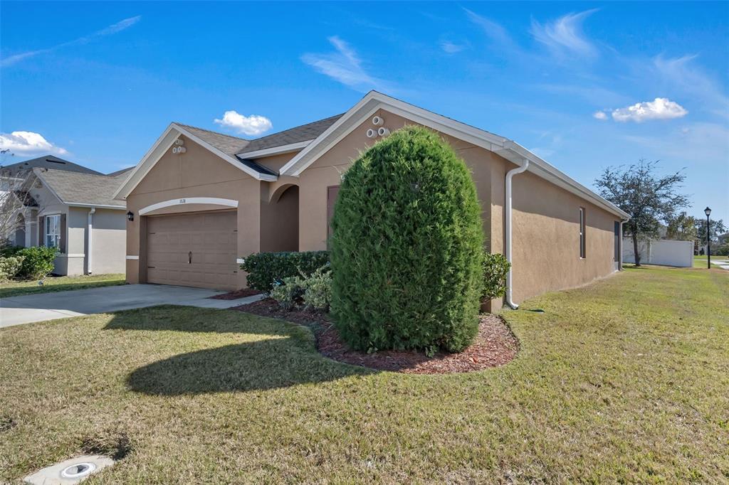 A single-story tan stucco house with a two-car garage and a large evergreen bush — 1828 PARTIN TERRACE ROAD, KISSIMMEE, FL
