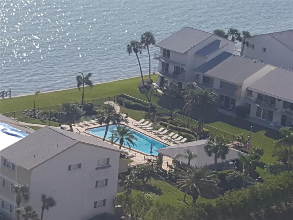 An aerial view shows a swimming pool surrounded by palm trees and multi-story residential buildings — 1301 GULF BOULEVARD UNIT 101, CLEARWATER BEACH, FL