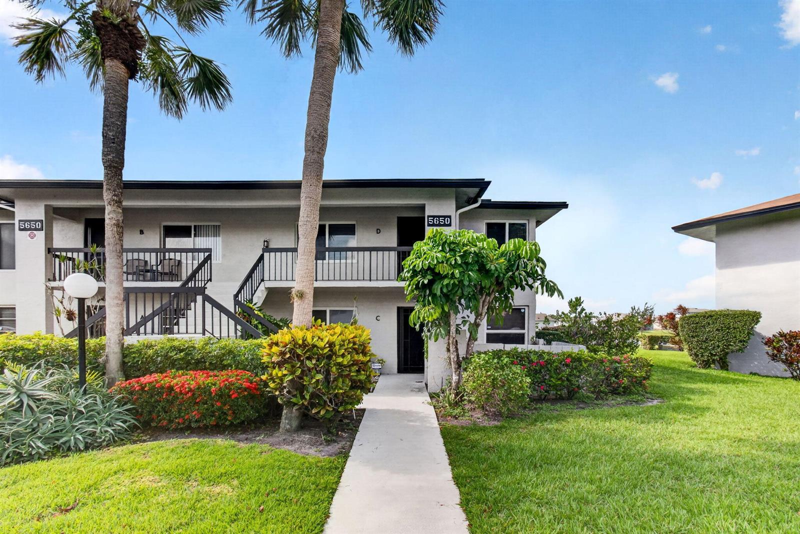 A two-story apartment building features a concrete walkway leading to the ground-floor entrance — 5650 Spindle Palm Court # D, Delray Beach FL 33484, DELRAY BEACH, FL