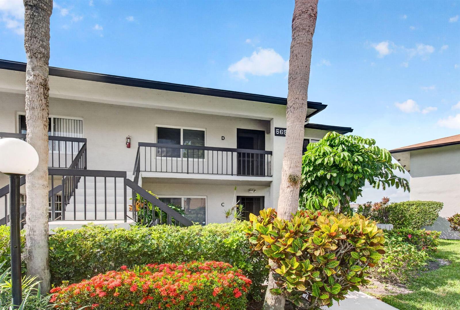 A two-story apartment building exterior features balconies, stairs, and landscaping under a blue sky — 5650 Spindle Palm Court # D, Delray Beach FL 33484, DELRAY BEACH, FL