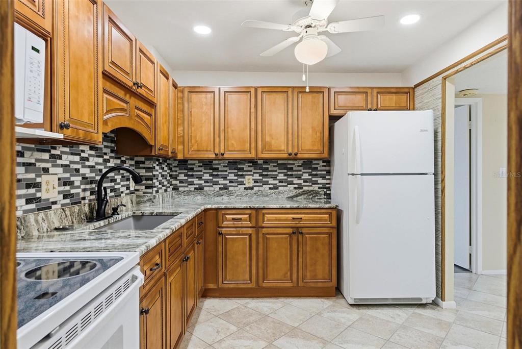 A kitchen with wood cabinets, granite countertops, a mosaic tile backsplash, and a white refrigerator — 925 N HALIFAX AVENUE UNIT 103, DAYTONA BEACH, FL