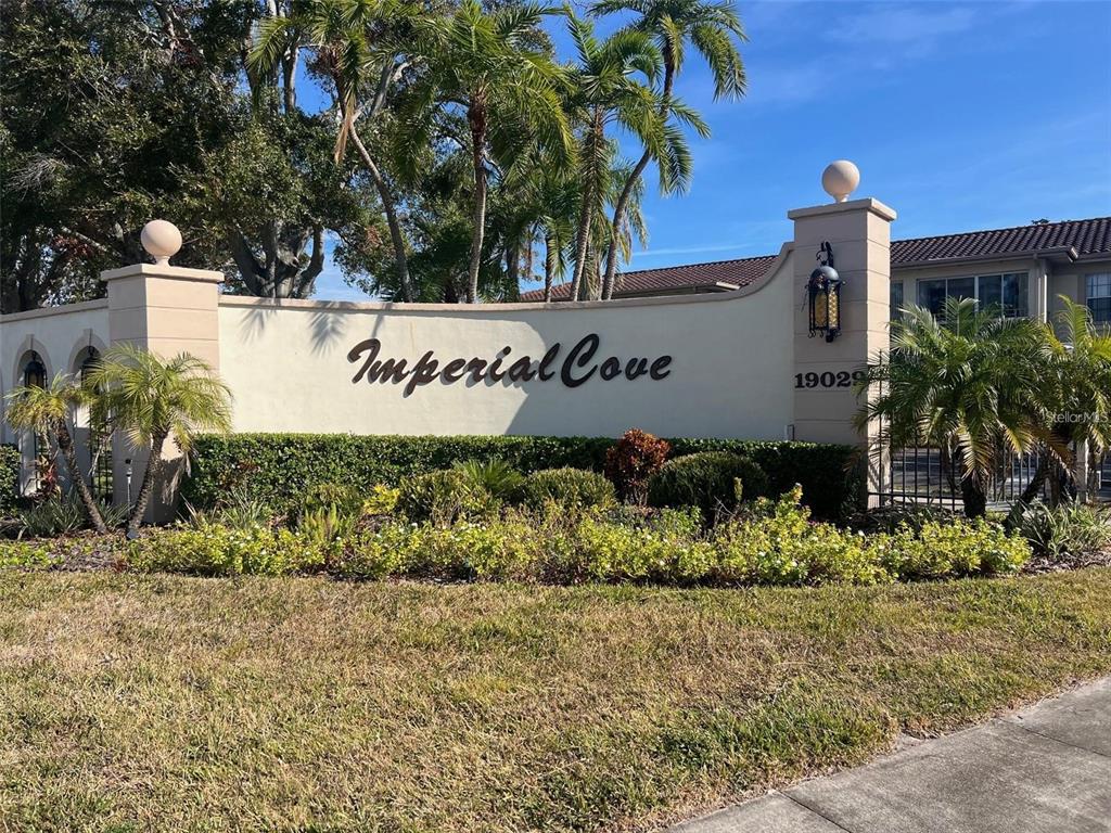 A beige stucco entrance sign for Imperial Cove features palm trees and landscaping — 19029 US HIGHWAY 19 N UNIT 6-14, CLEARWATER, FL