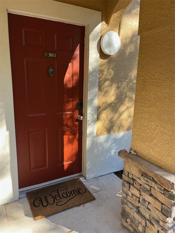 A red front door with a welcome mat is set into a stucco and stone exterior — 2107 PORTOFINO PLACE UNIT 3011, PALM HARBOR, FL