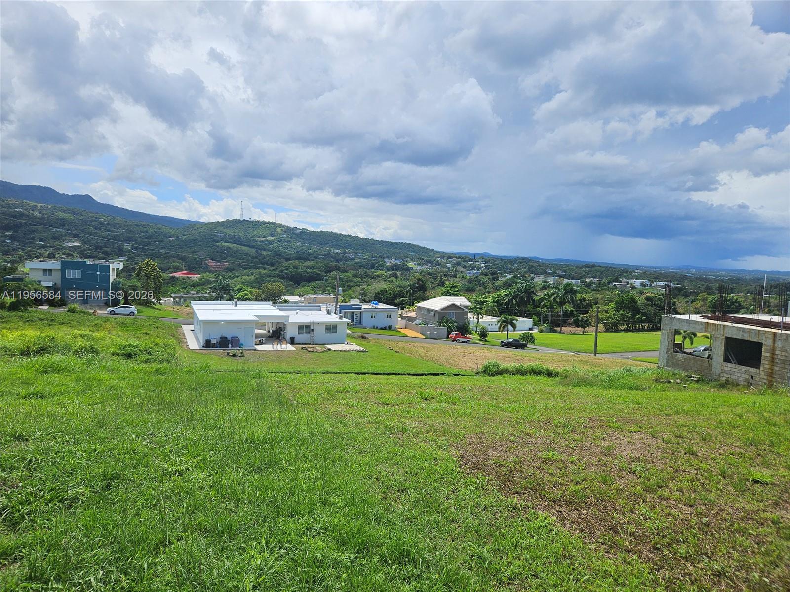 A grassy, sloping lot overlooks a rural landscape with several houses under a cloudy sky — URB MIRADOR DE PALMER . SOLAR 41 RIO GRANDE, PR 00745, PR
