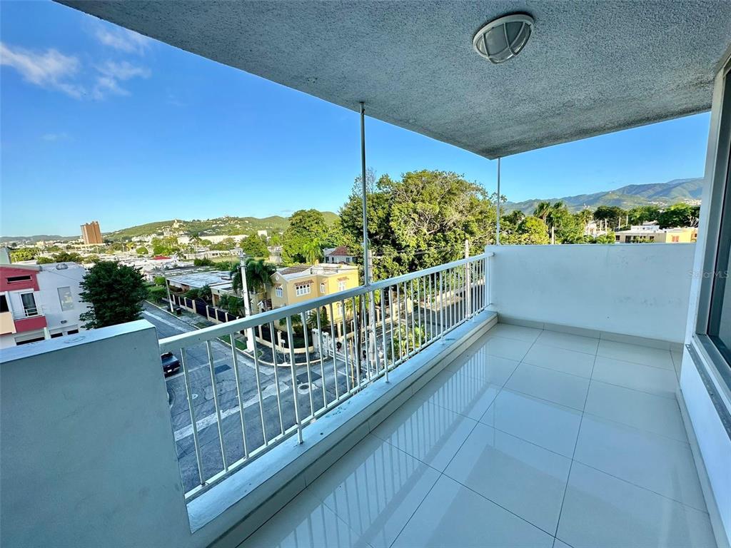 A tiled balcony with a white metal railing overlooks a city street and mountains — 1802 ALCAZAR UNIT 404, PONCE, PR