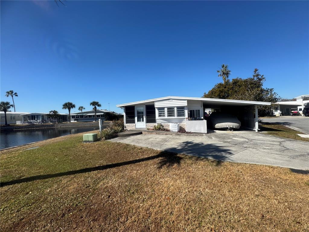A single-story mobile home with a carport sits beside a canal under a clear sky — 4842 BRITNI WAY UNIT 058, ZEPHYRHILLS, FL