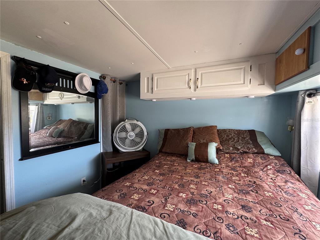 A bedroom features a bed with patterned bedding, a wall-mounted mirror, and overhead white cabinets — 4842 BRITNI WAY UNIT 058, ZEPHYRHILLS, FL