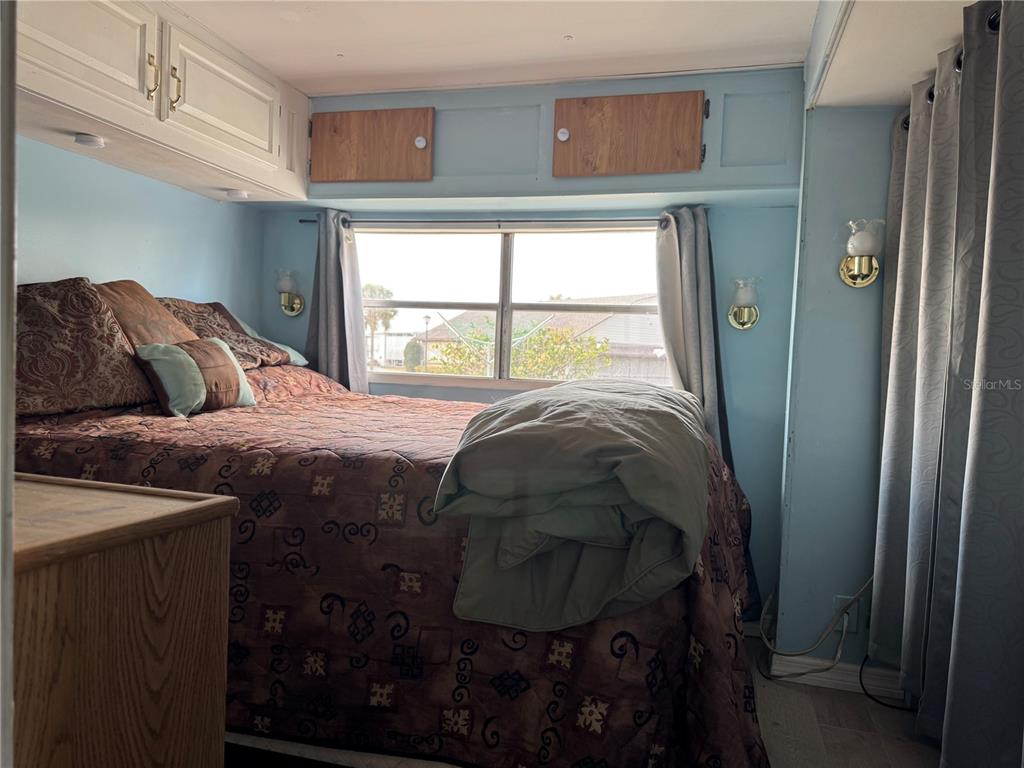 A bedroom features a bed with brown bedding, light blue walls, and overhead wooden cabinets — 4842 BRITNI WAY UNIT 058, ZEPHYRHILLS, FL