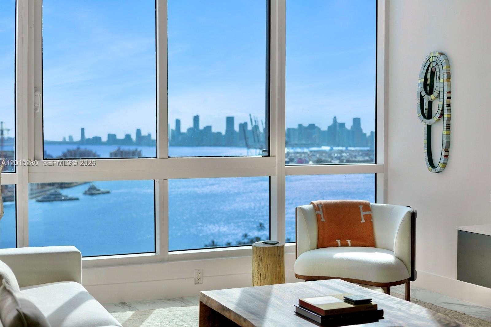 A modern living room features floor-to-ceiling windows overlooking a bay and a city skyline — 100 S Pointe Dr # 2003, Miami Beach FL 33139, MIAMI BEACH, FL