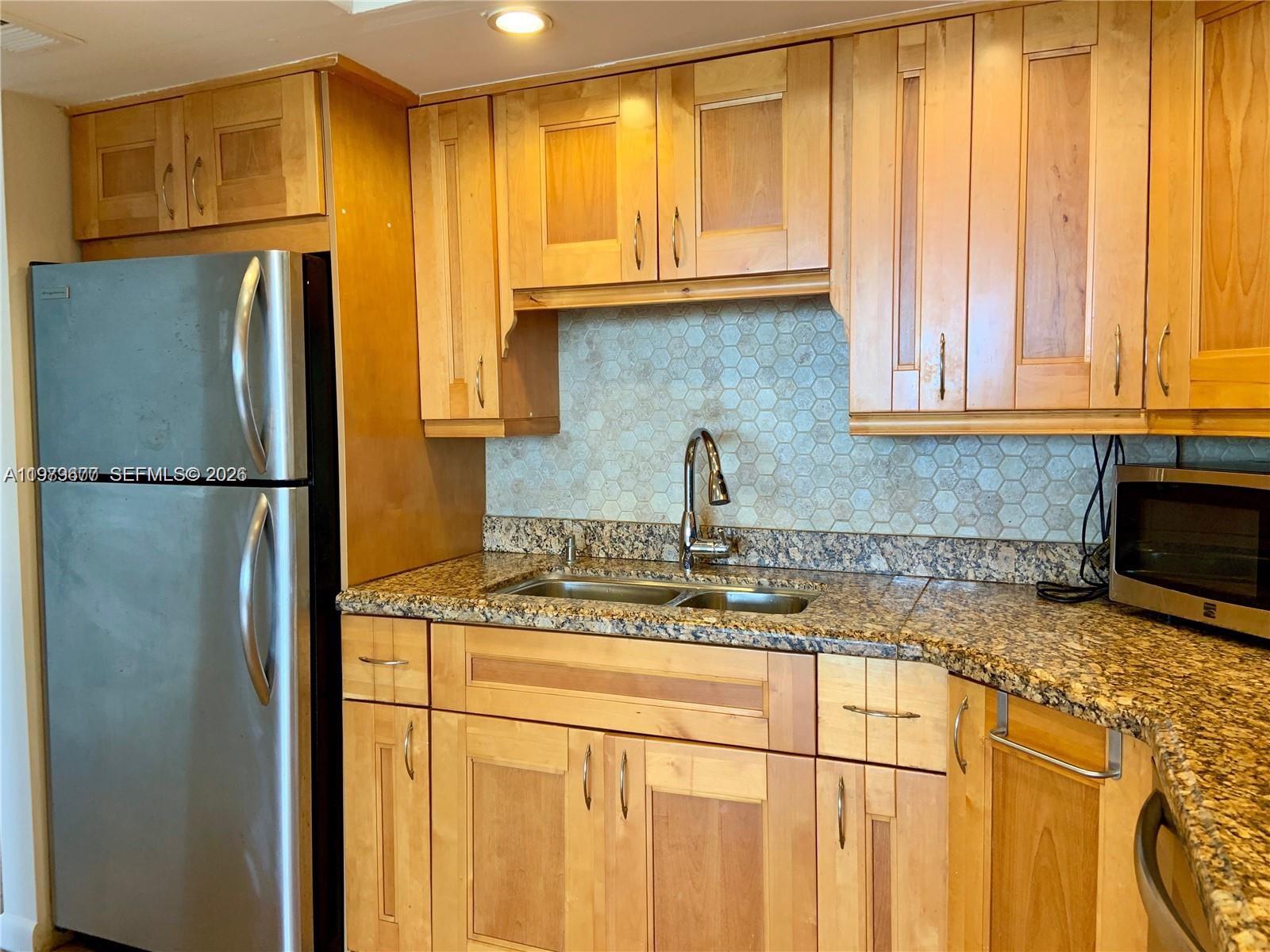 A kitchen features light wood cabinets, granite countertops, a stainless steel refrigerator, and hexagonal backsplash — 2025 NE 164th St # 1015, North Miami Beach FL 33162, NORTH MIAMI BEACH, FL