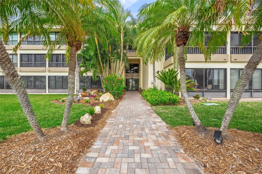 A brick walkway leads to the entrance of a multi-story apartment building with palm trees — 101 WHISPERING SANDS DRIVE UNIT 105, SARASOTA, FL