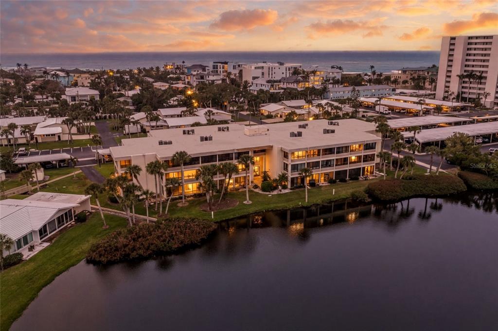An aerial view of a multi-story apartment building situated next to a pond at sunset — 101 WHISPERING SANDS DRIVE UNIT 105, SARASOTA, FL