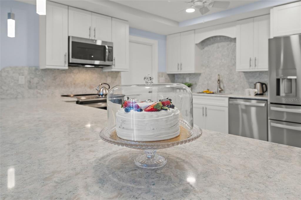 A white kitchen features a cake on a stand atop a marble-topped island — 101 WHISPERING SANDS DRIVE UNIT 105, SARASOTA, FL
