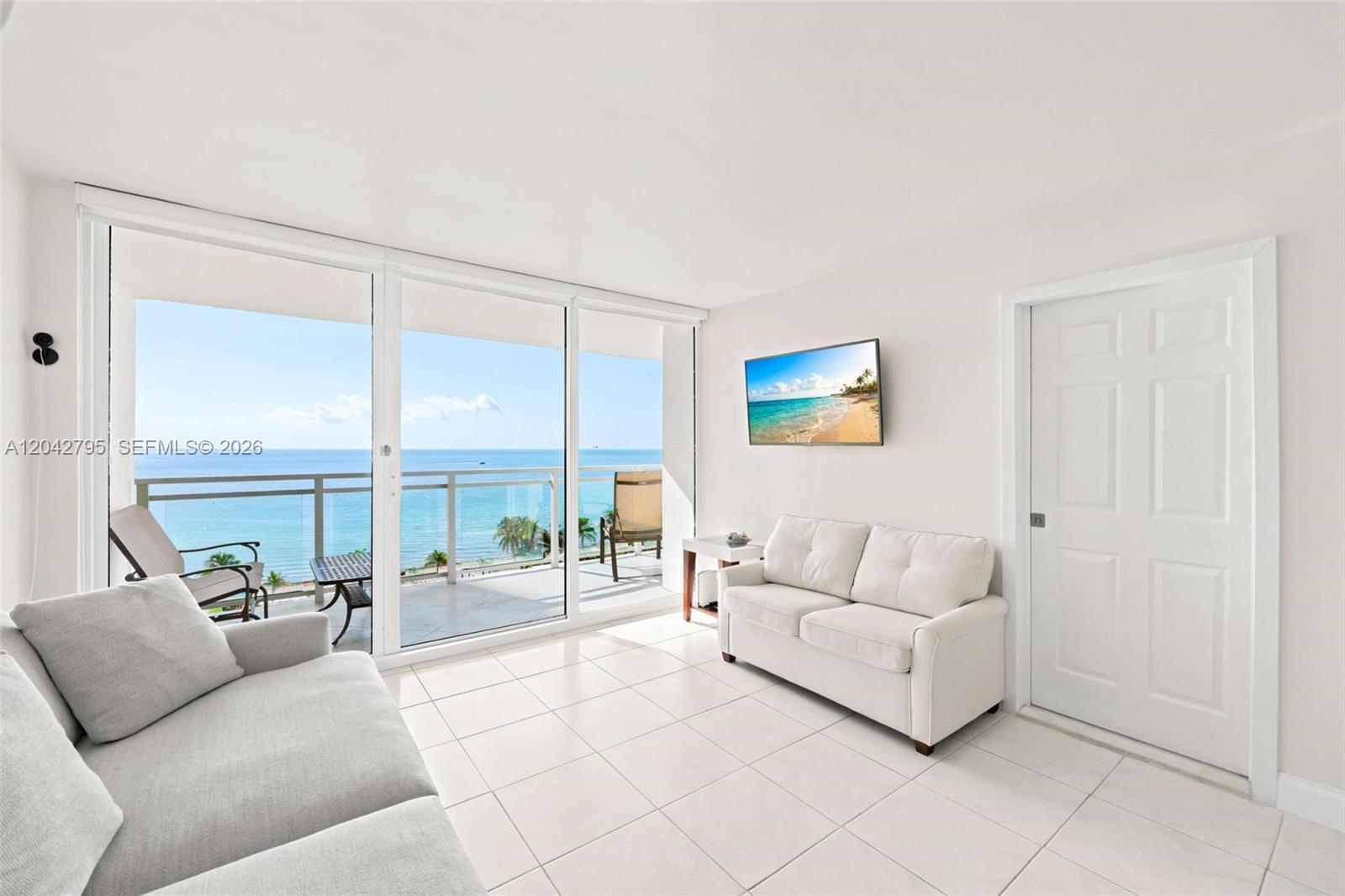 A living room with white tile floors, two sofas, a wall-mounted television, and a balcony — 2030 S Ocean Dr # 816, Hallandale Beach FL 33009, HALLANDALE BEACH, FL