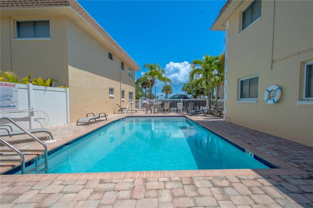 A rectangular swimming pool sits between two beige multi-story buildings on a paved patio — 9050 BLIND PASS ROAD UNIT 9, ST PETE BEACH, FL