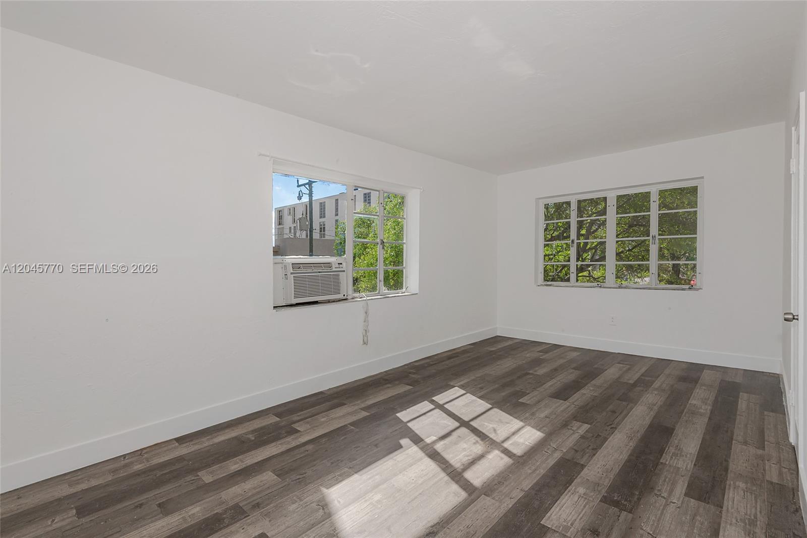 An empty room features white walls, dark wood-look flooring, and two windows with exterior views — 8340 Crespi Blvd # 20, Miami Beach FL 33141, MIAMI BEACH, FL