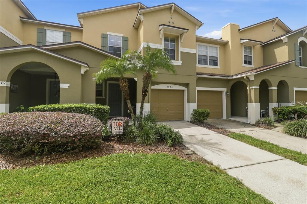 A two-story townhouse exterior features tan and olive siding, two garage doors, and landscaping — 3845 GLIDING PLACE, SANFORD, FL
