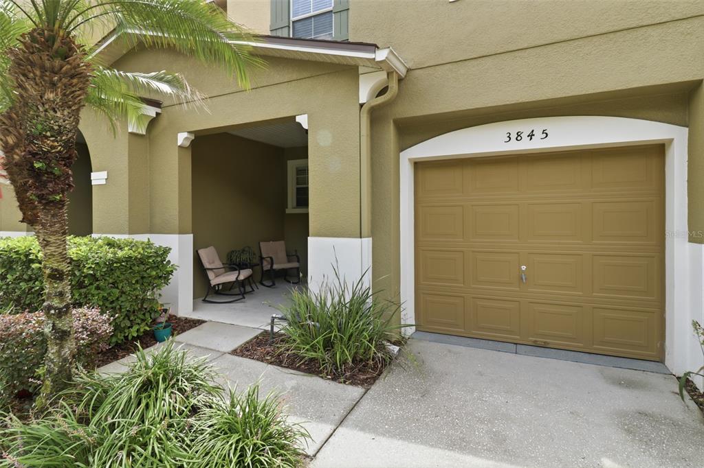 A tan exterior home features a covered porch with two chairs and a garage door — 3845 GLIDING PLACE, SANFORD, FL