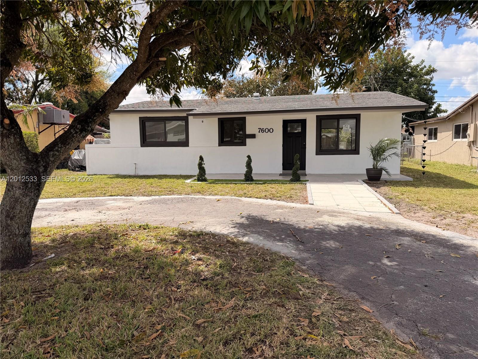A single-story white house with dark trim and a paved walkway sits behind a tree — 7600 Venetian St, Miramar FL 33023, MIRAMAR, FL