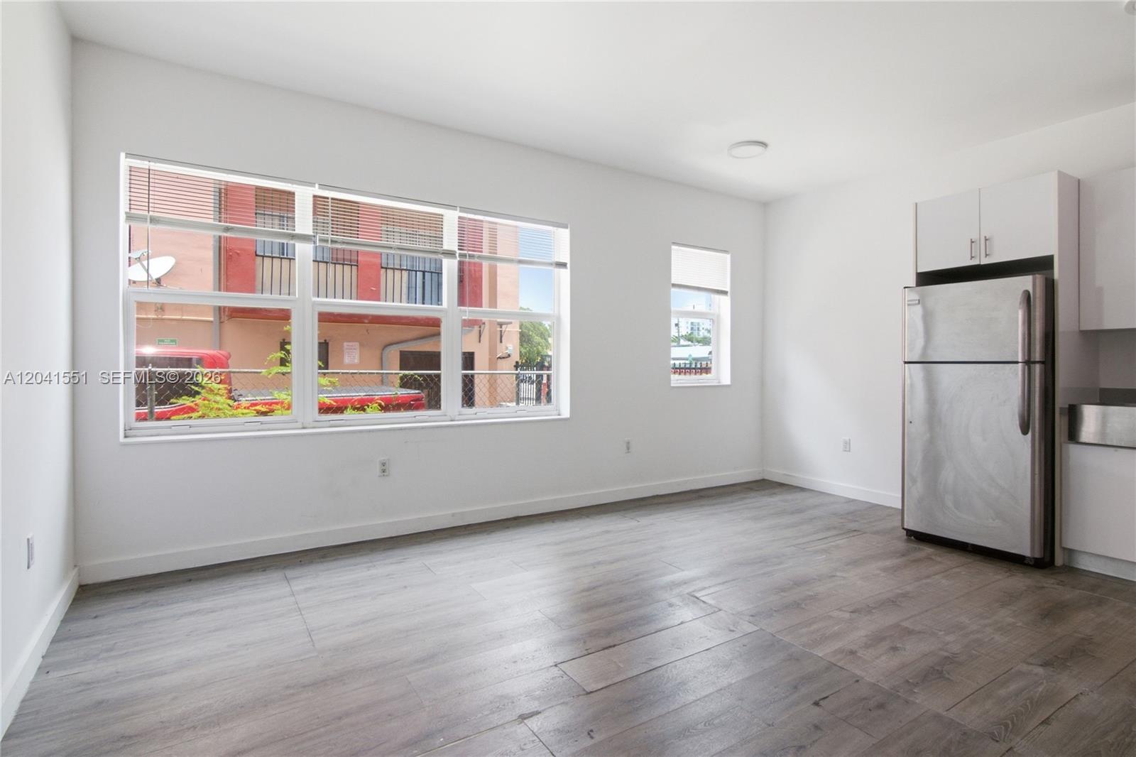 A bright, unfurnished room features wood-look flooring, white walls, a large window, and a refrigerator — 402 NW 12th Ave # 111, Miami FL 33128, MIAMI, FL
