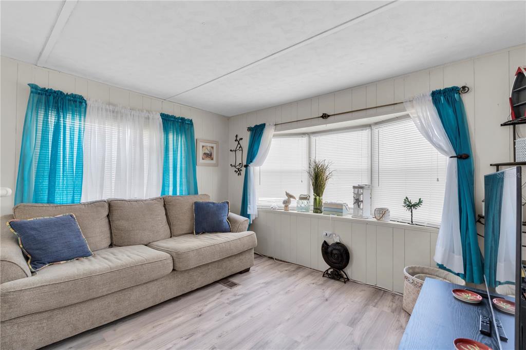 A living room features a beige sofa, light wood-look flooring, and windows with blue curtains — 7801 34TH AVENUE N UNIT 27, ST PETERSBURG, FL