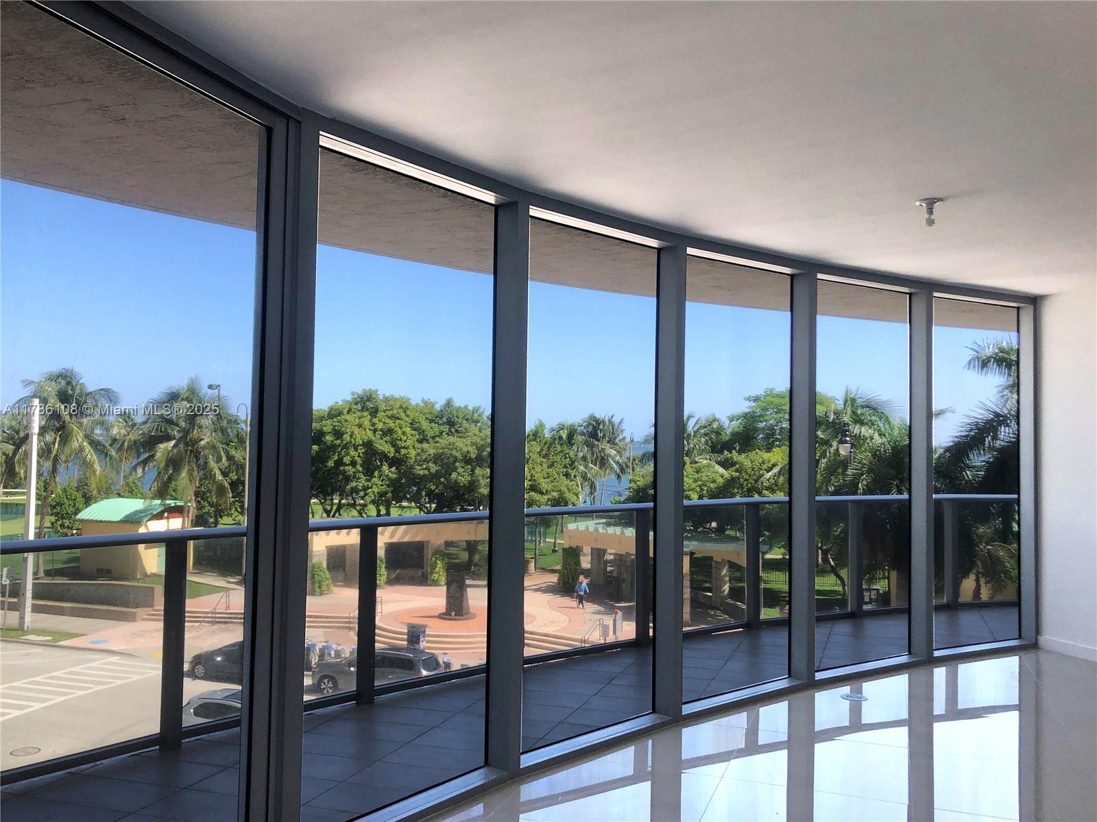 A room with floor-to-ceiling windows overlooking a park, palm trees, and a paved plaza — 488 NE 18th St # 215, Miami FL 33132, MIAMI, FL