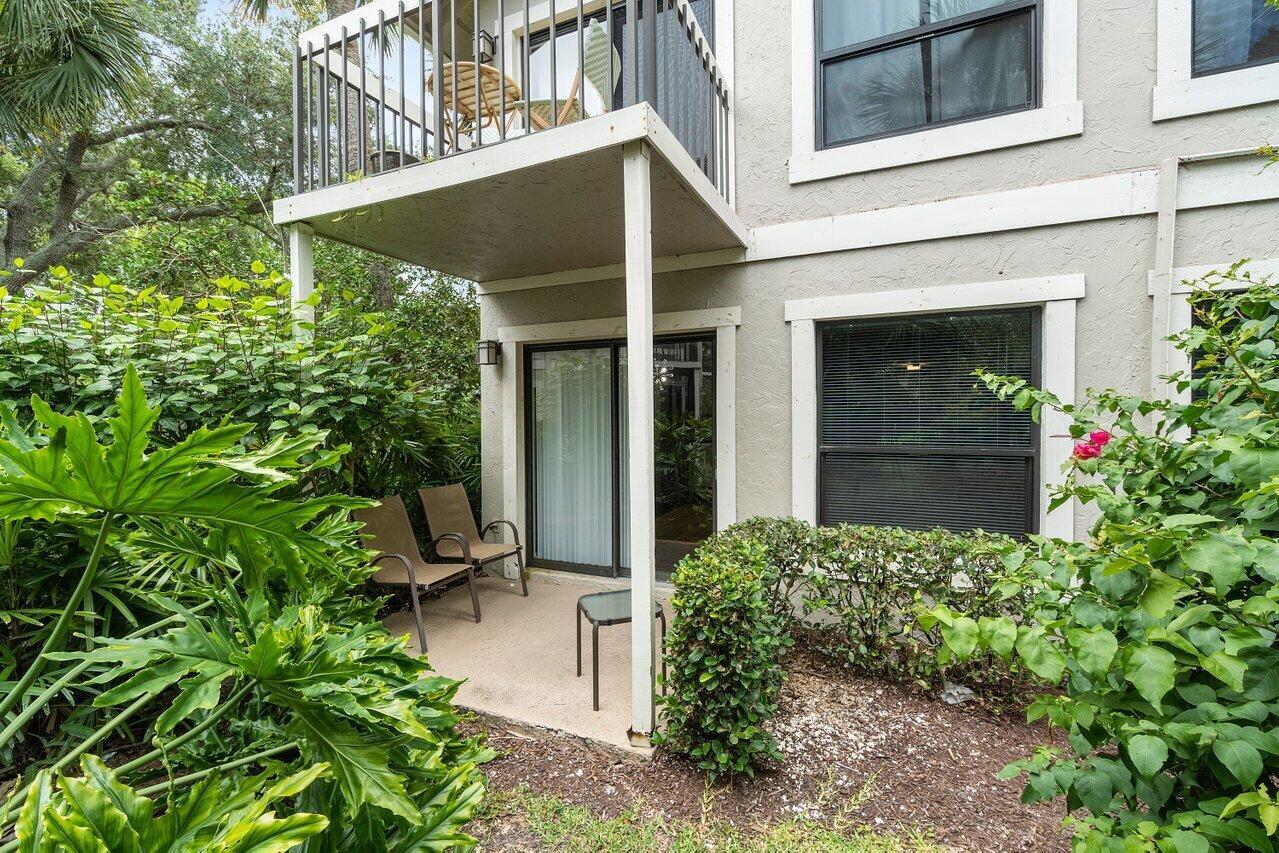 A ground-level patio with two chairs sits beneath a second-story balcony surrounded by lush landscaping — 11863 Wimbledon Circle # 407, Wellington FL 33414, WELLINGTON, FL